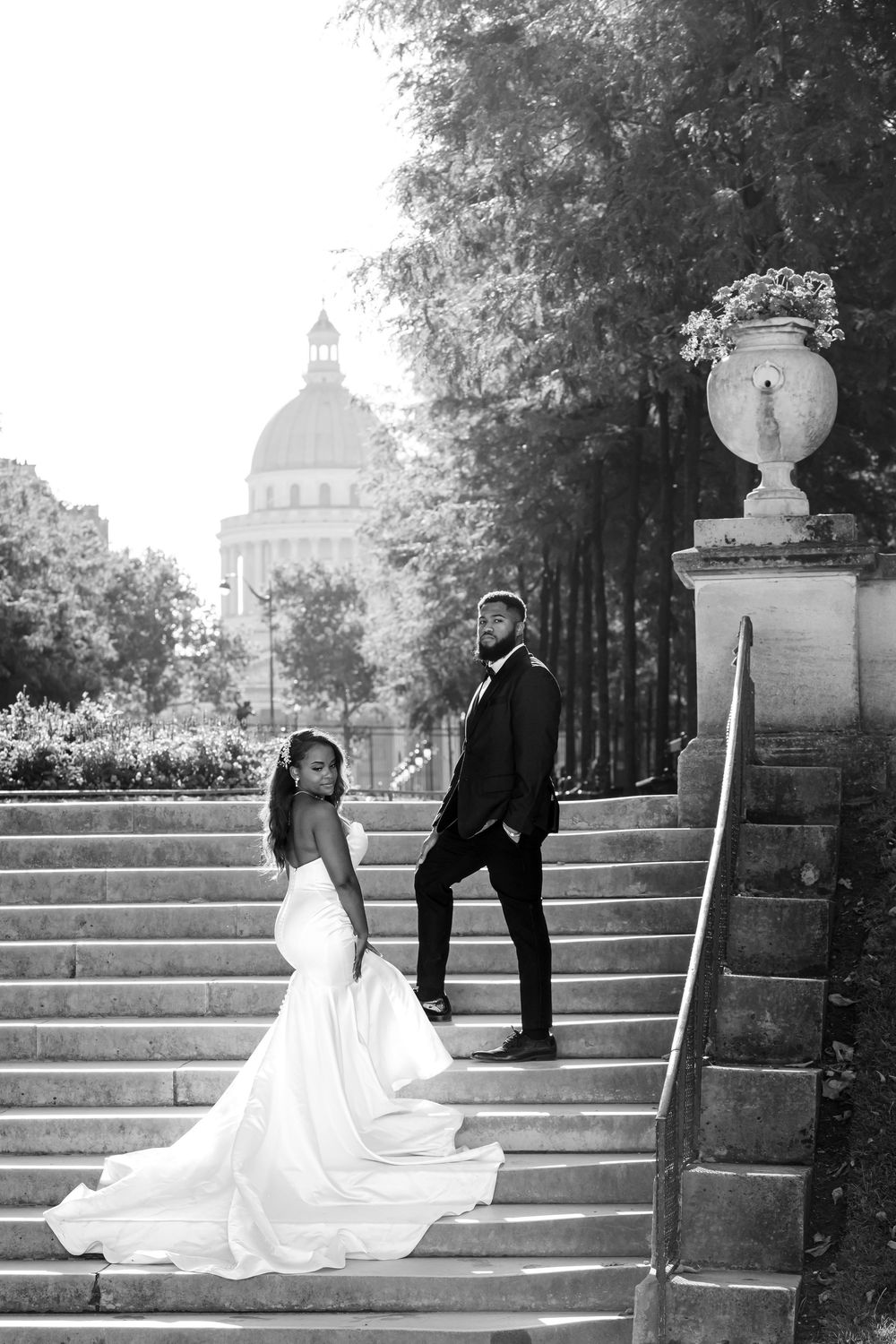 A bride and groom pose elegantly on stone steps with a domed building and ornate garden urn in the background.