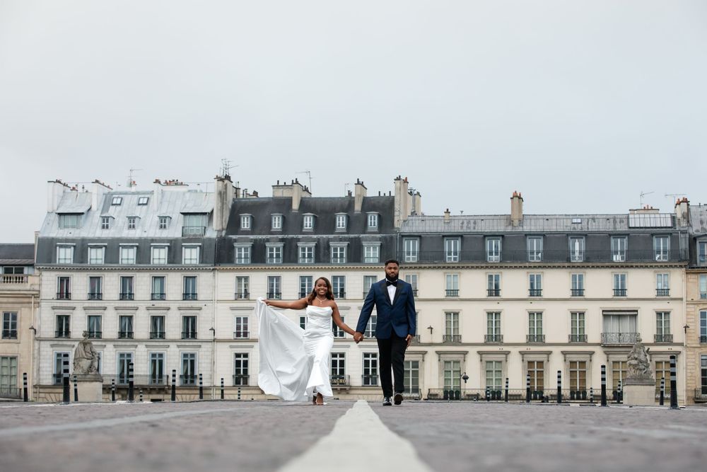 A couple in wedding attire walks along a scenic balcony with classic Parisian architecture in the background.