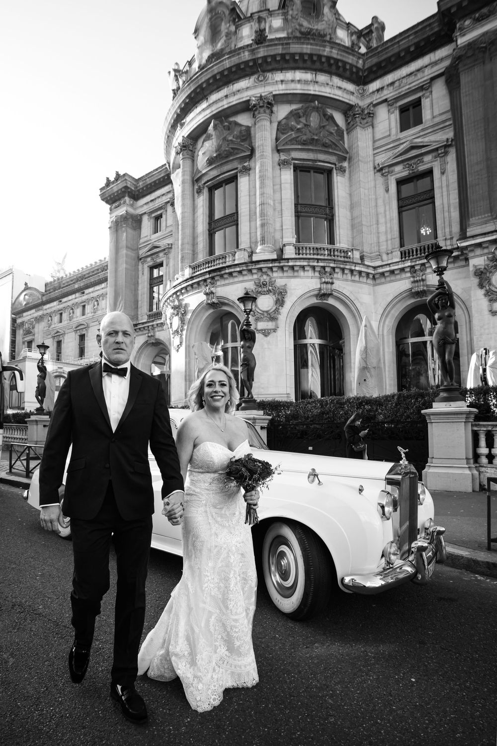 Black and white wedding photo series showing a couple walking with a classic vintage car outside an ornate building.