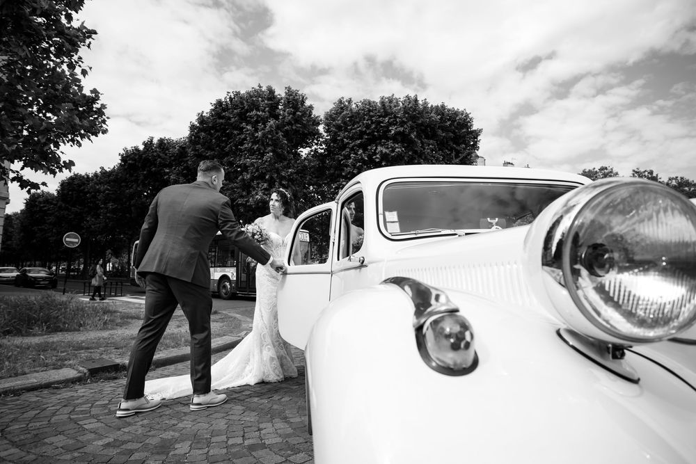 Black and white photo of a classic vintage car at an outdoor wedding with couple entering the vehicle.
