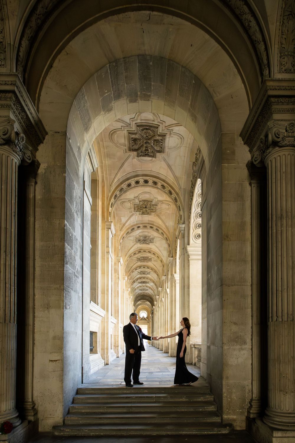 Bride and groom stand in silhouette within a grand arched colonnade hallway with classical Louvre architecture and natural lighting.