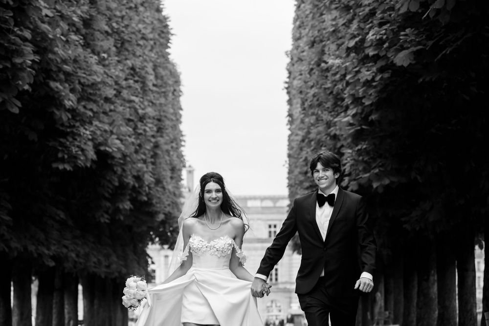 A bride and groom walk hand in hand through a garden path lined with tall manicured hedges on their wedding day.