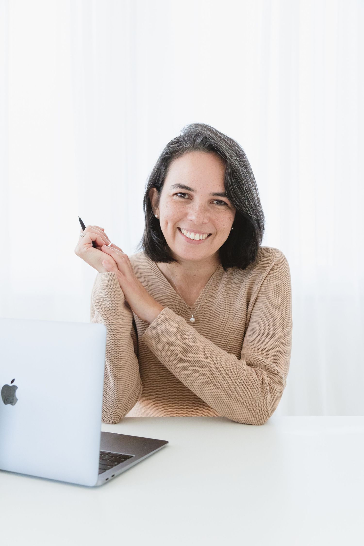 A smiling professional works at a desk with a MacBook laptop in a bright, minimalist setting.