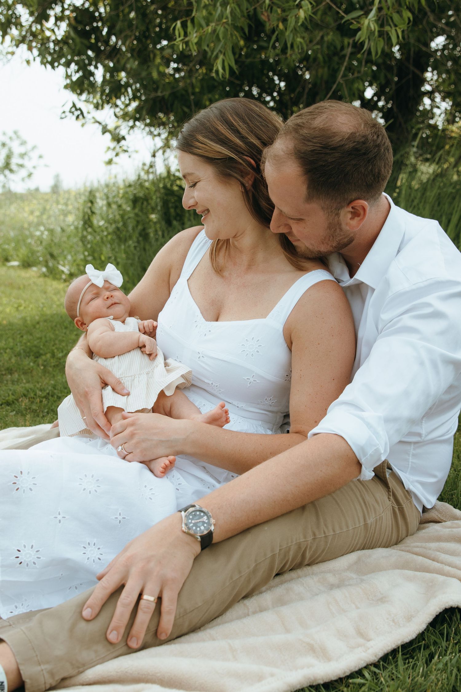 Intimate family moment captured on white blanket in sunny outdoor setting.