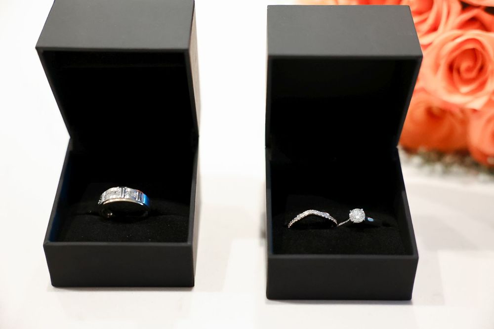 Two engagement rings displayed in black jewelry boxes against a coral rose background.