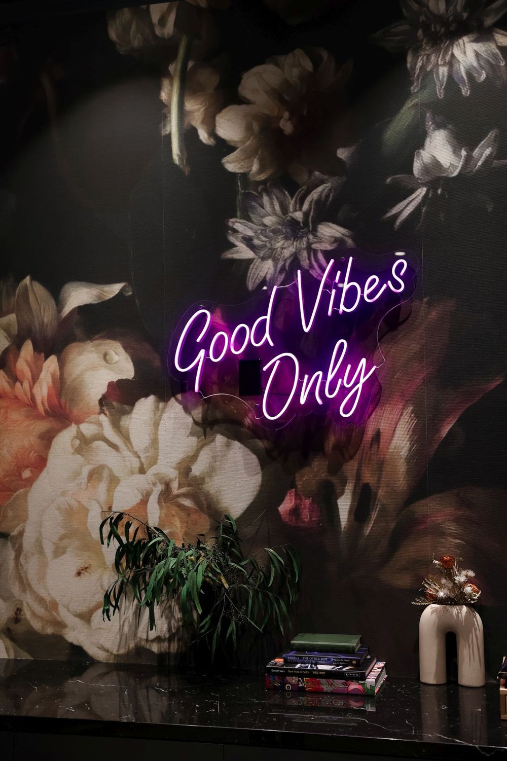 A dark moody wall with floral wallpaper features a purple neon sign reading 'Good Vibes Only' above books and decor.