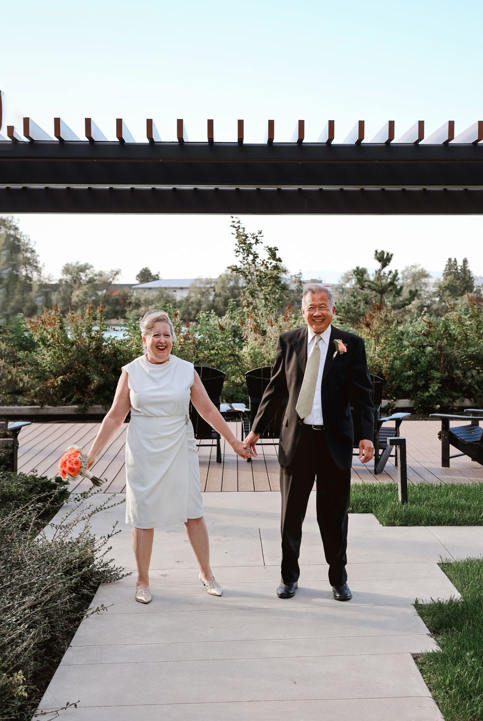 A couple walks hand in hand on an outdoor path under a wooden pergola.