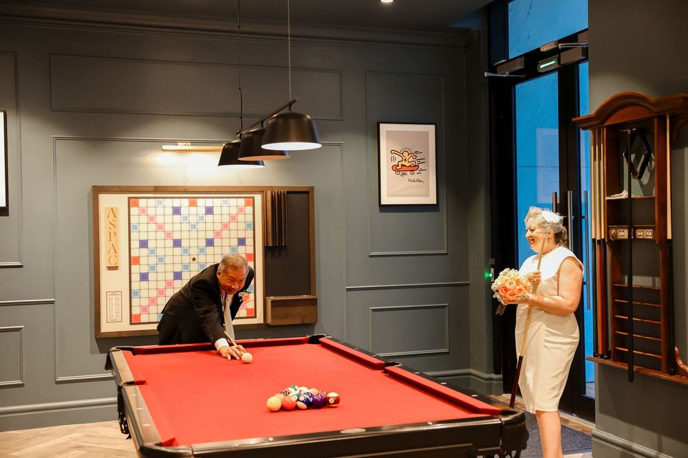 Two people play pool in a stylish grey room with pendant lighting.