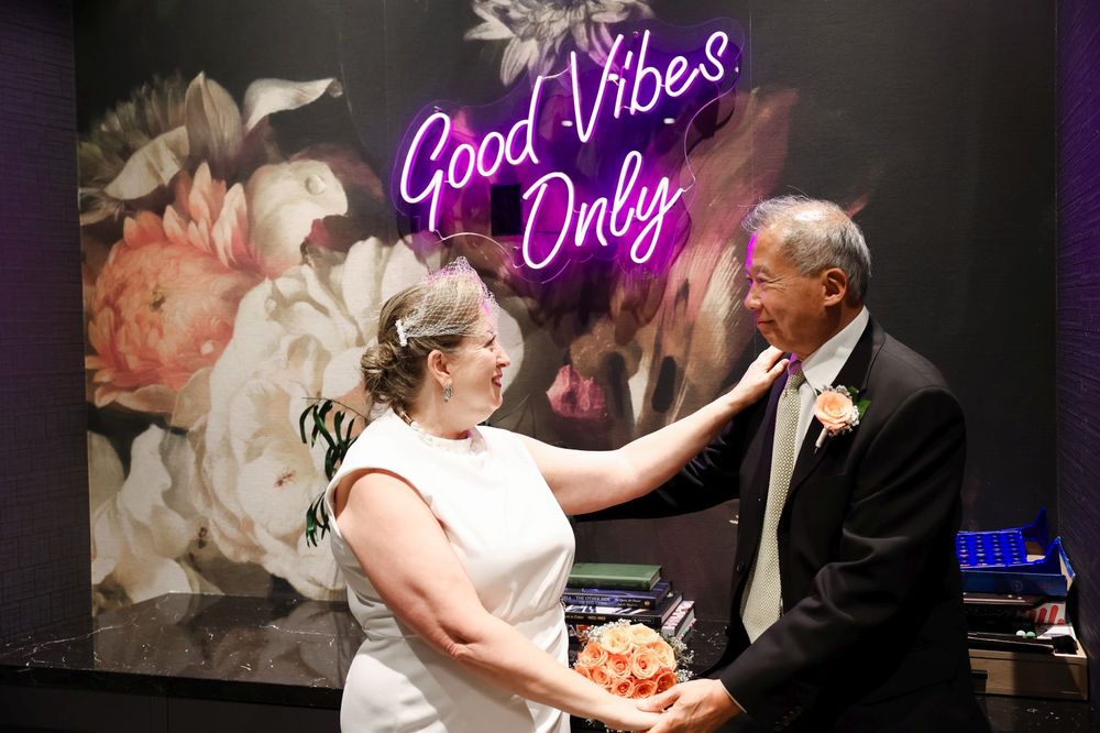 A couple dances together in front of a floral wall with neon 'Good Vibes Only' sign.