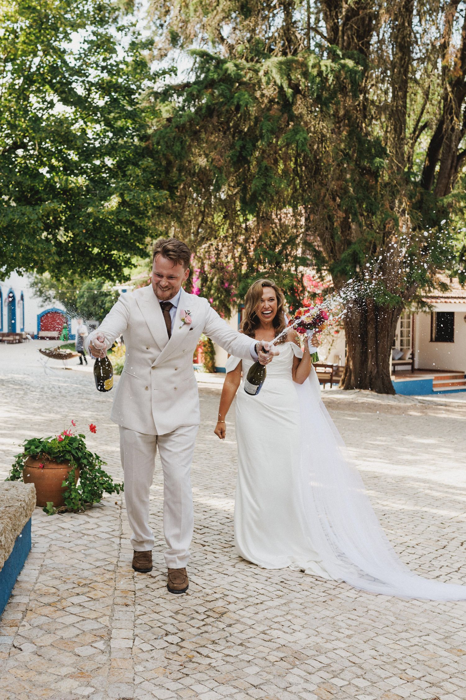 A couple in white wedding attire joyfully walk together on a cobblestone street under overhanging trees.