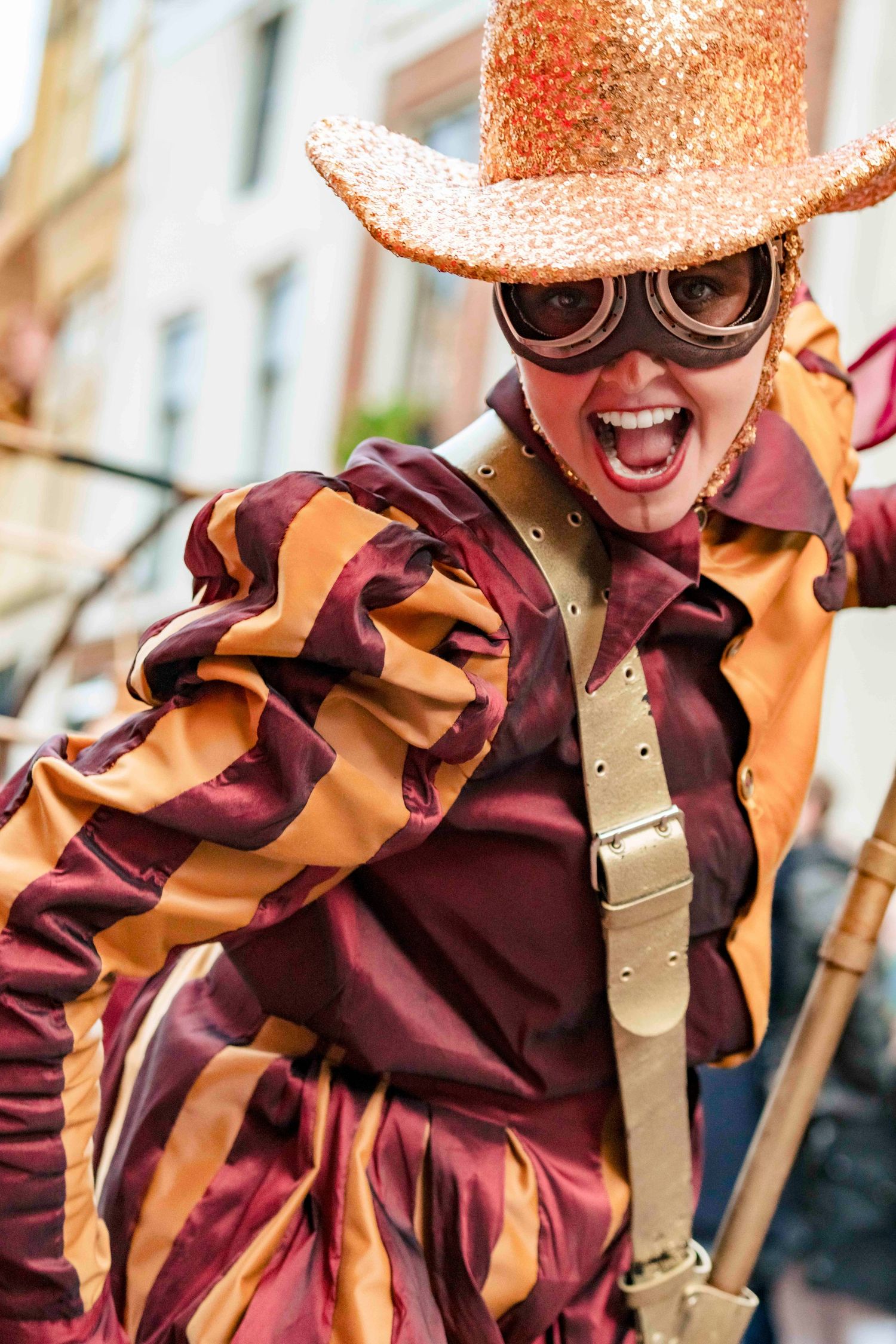 A performer in a sparkly orange hat and striped burgundy costume expresses joy during a theatrical performance.