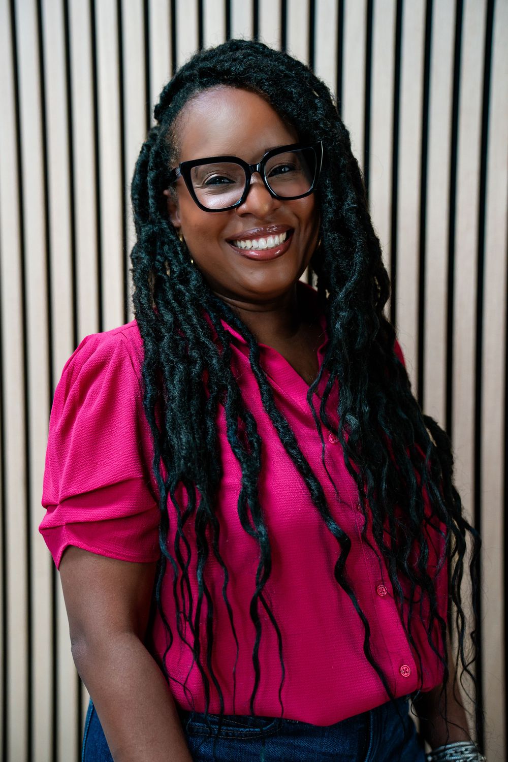 Smiling woman with long locs and black glasses wearing a pink top, standing against a striped wall.