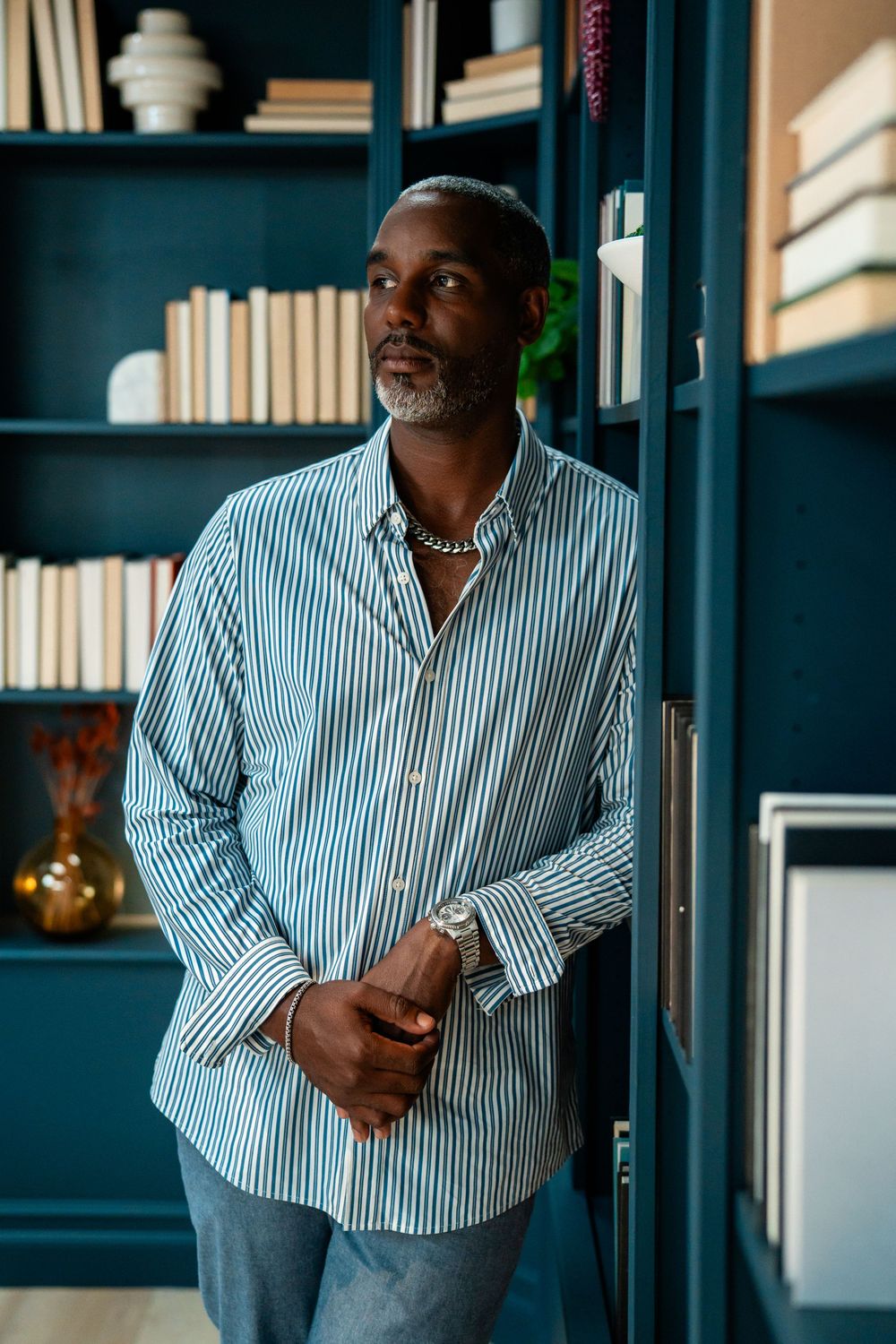 A stylish man in a striped shirt leans against a teal bookshelf, wearing a chain necklace in a modern library setting.