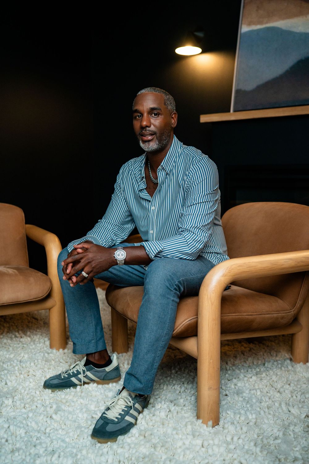 A man in a striped shirt and jeans sits relaxed in a tan leather chair, wearing sneakers and a watch.