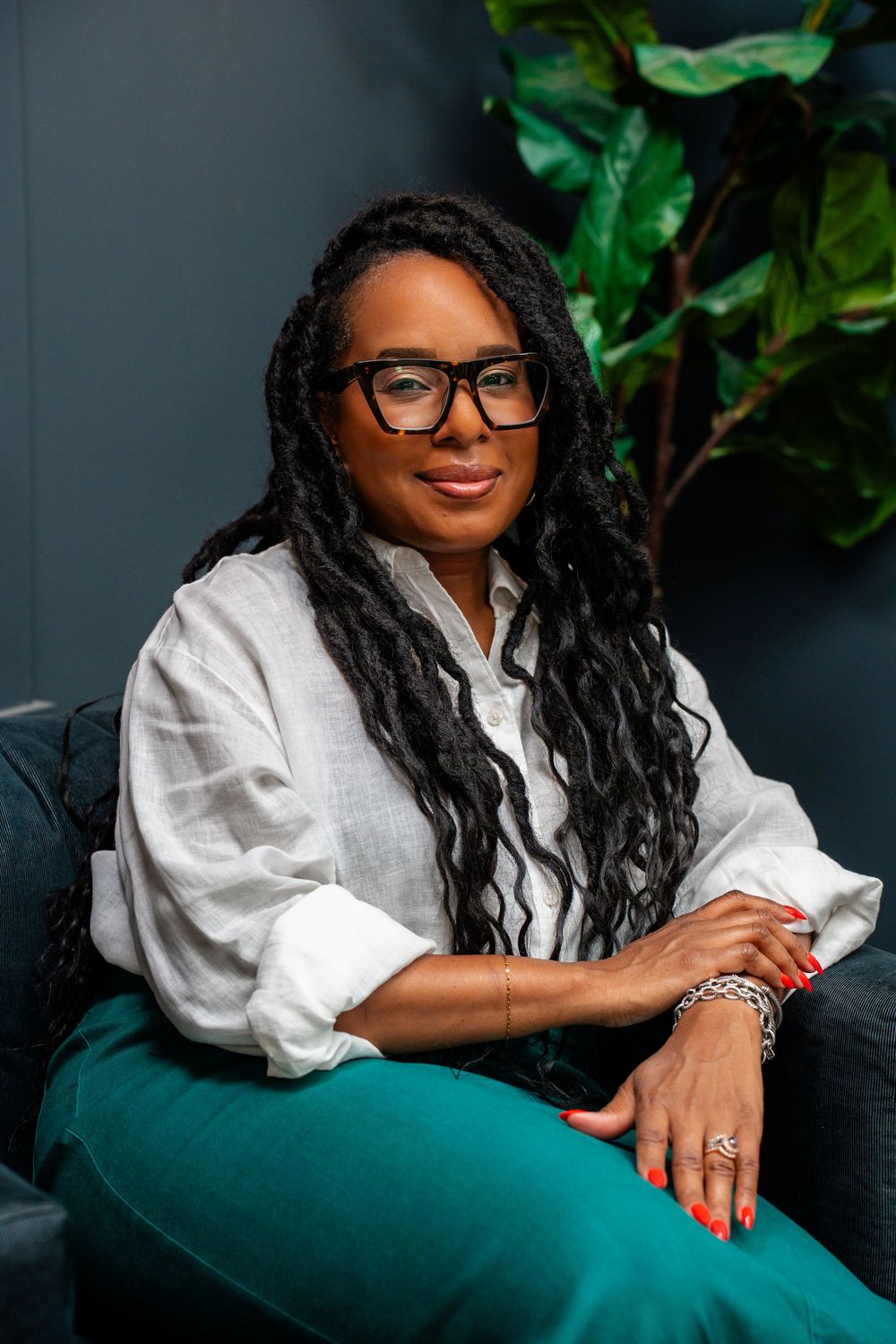 A woman with long braids and glasses sits confidently in a chair, wearing a white blouse and teal pants.