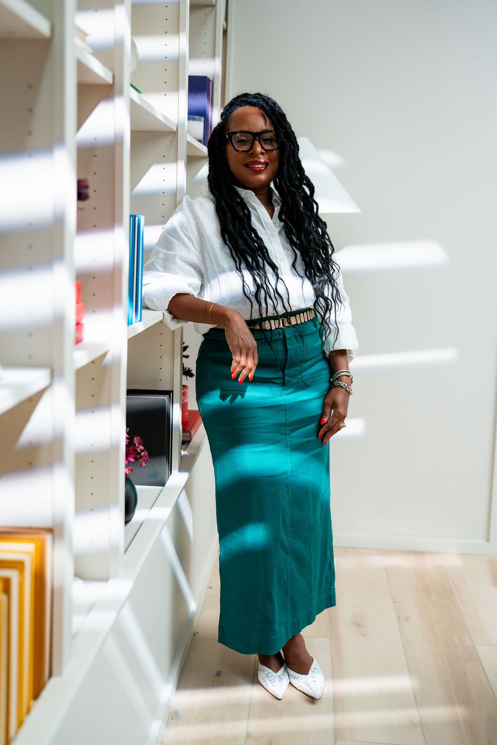A stylish woman in a white shirt and teal maxi skirt poses beside white shelves in a bright modern space.