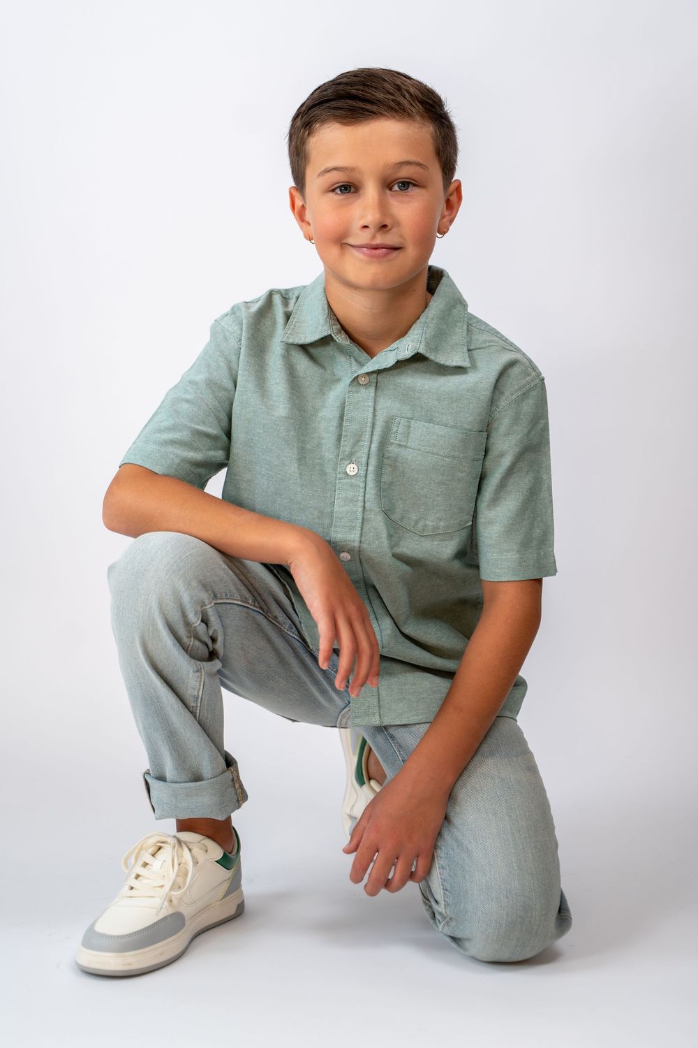 Young person in a mint green button-down shirt and light jeans kneels in a casual pose against a white backdrop.