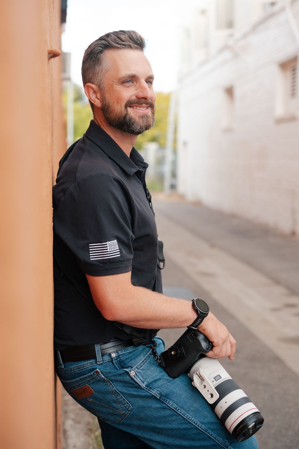 Professional photographer in black polo and jeans holding a camera with telephoto lens while smiling on urban street.
