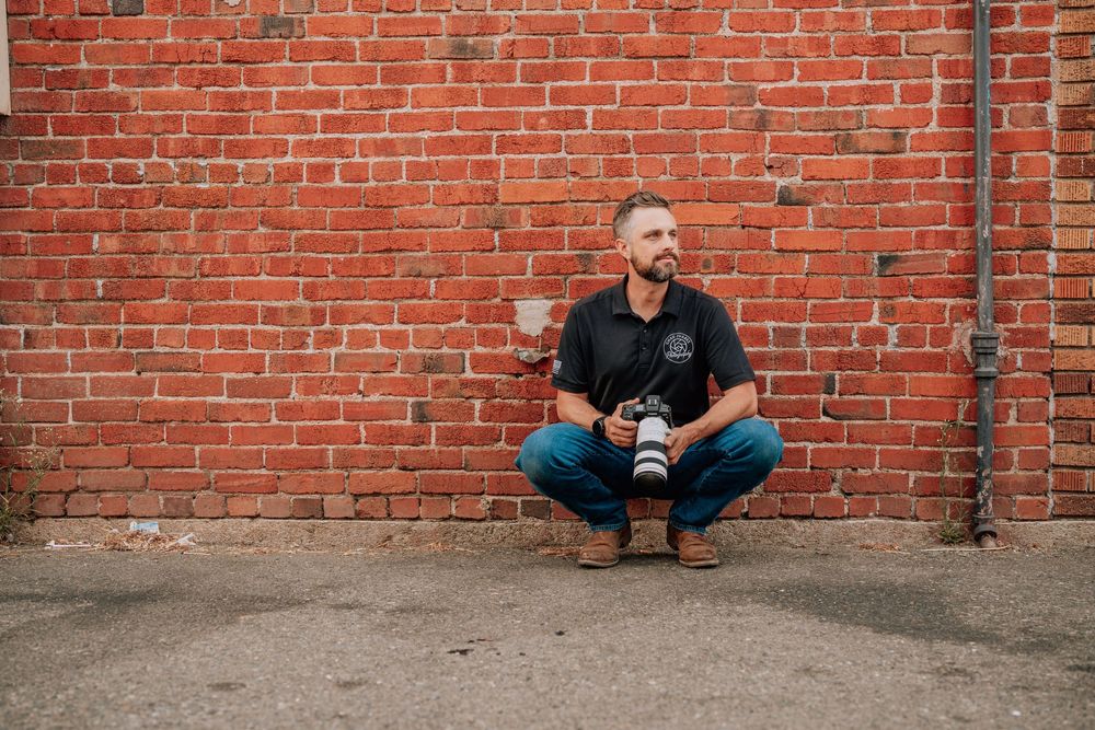 A person in dark clothing crouches against a weathered red brick wall while holding a beverage.