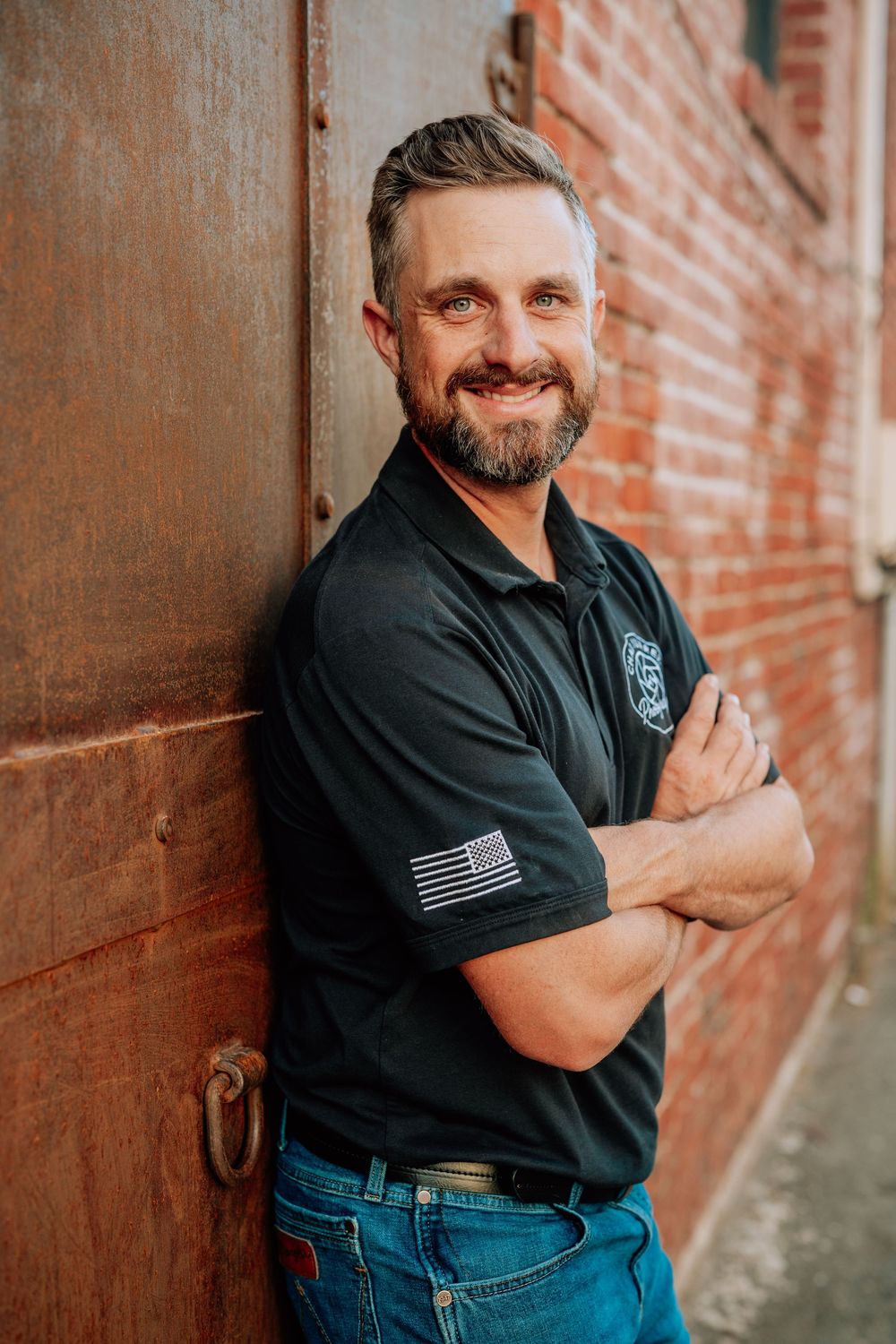 Someone in a black polo shirt with an American flag patch poses confidently against a rustic brick wall.
