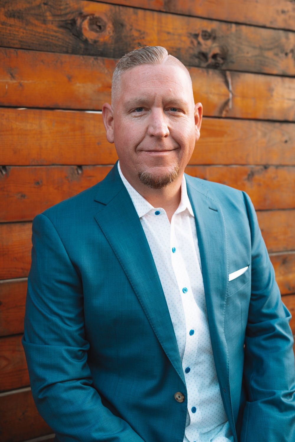 Professional headshot of man in teal blazer against wooden wall background.