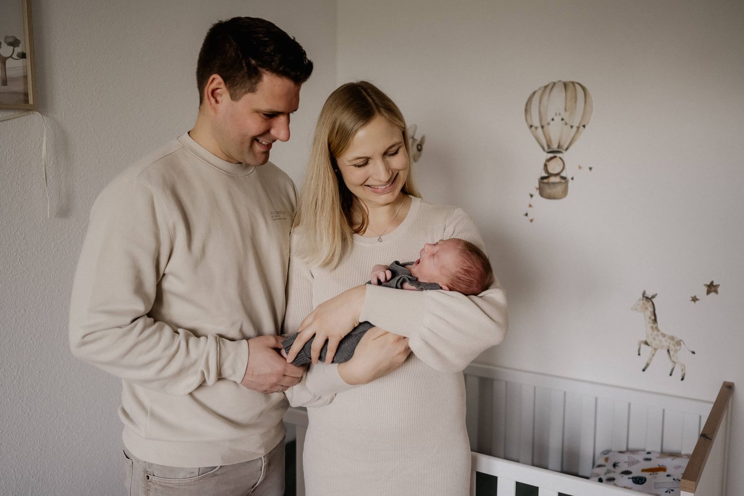 Parents lovingly gaze at their newborn baby in a cozy nursery room with beige neutral decor.