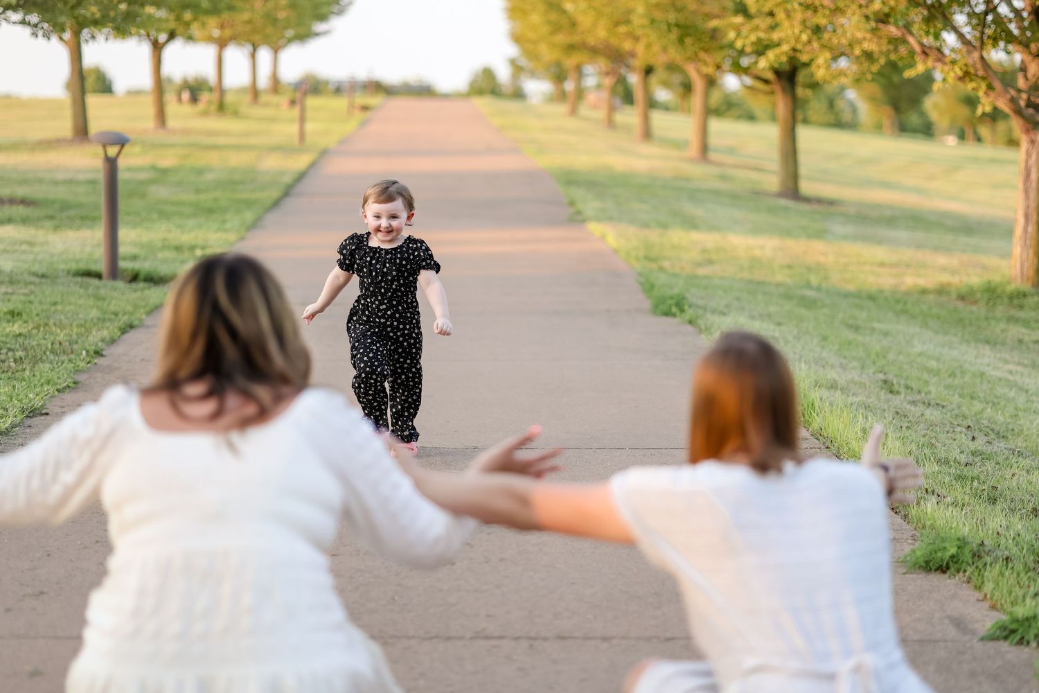 Child in black outfit running towards two people with arms outstretched on tree-lined path.