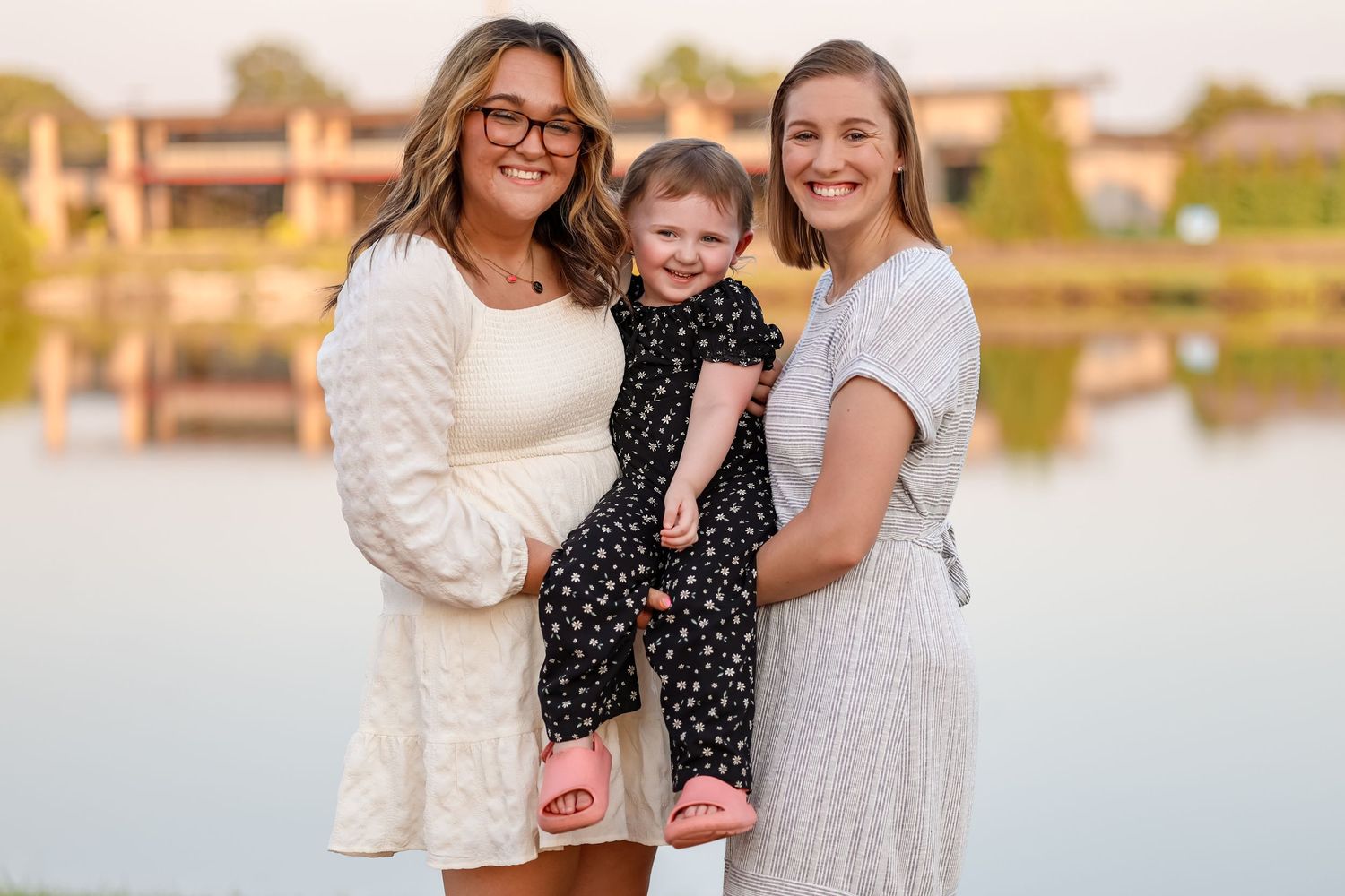 Three people posing together at sunset by a lake wearing white and polka dot dresses.