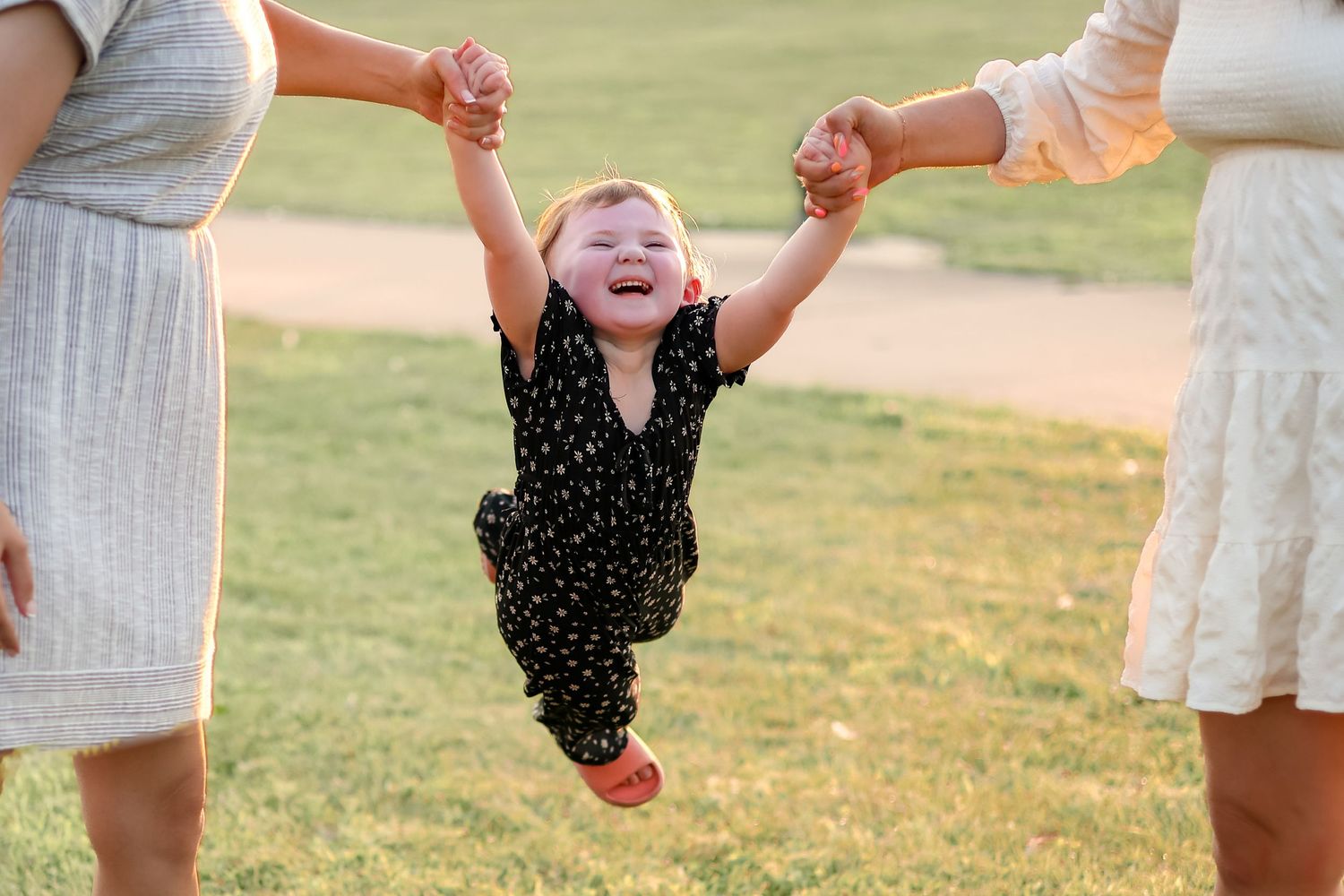 Child in polka dot outfit swinging joyfully between two adults on path.