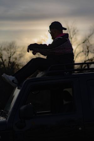 A silhouette of someone lying on top of a car's roof against a dramatic dusk sky.