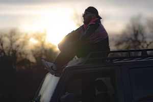 A silhouetted figure sits atop a vehicle at sunset while trees are visible in the background.