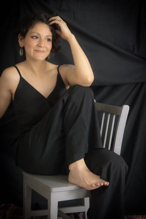 Woman in black jumpsuit sitting barefoot on white chair against dark backdrop, smiling with hand in hair.