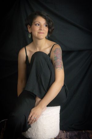Young woman with short dark hair and rose tattoo on arm, wearing black jumpsuit, seated against dark backdrop.