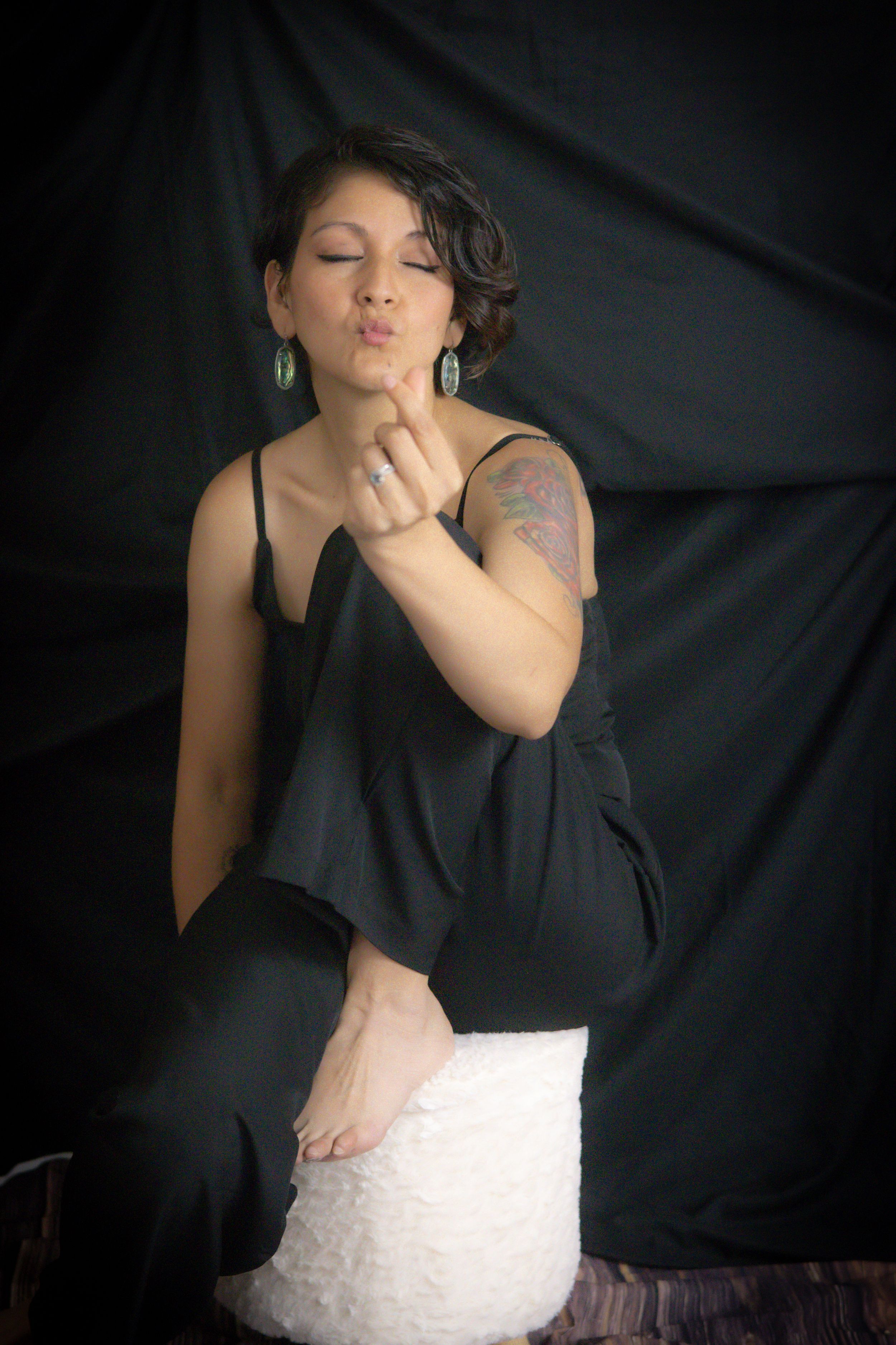 Woman in black outfit blowing a kiss, showing tattoo on arm, wearing teal earrings, seated on white stool.