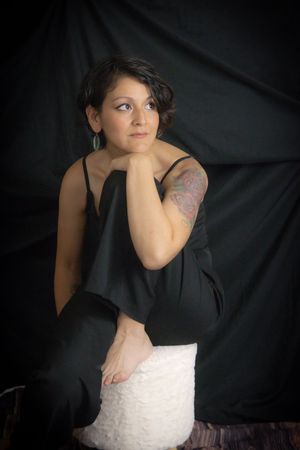 Woman in black jumpsuit sitting on white stool with tattoo on arm, posing against dark backdrop.