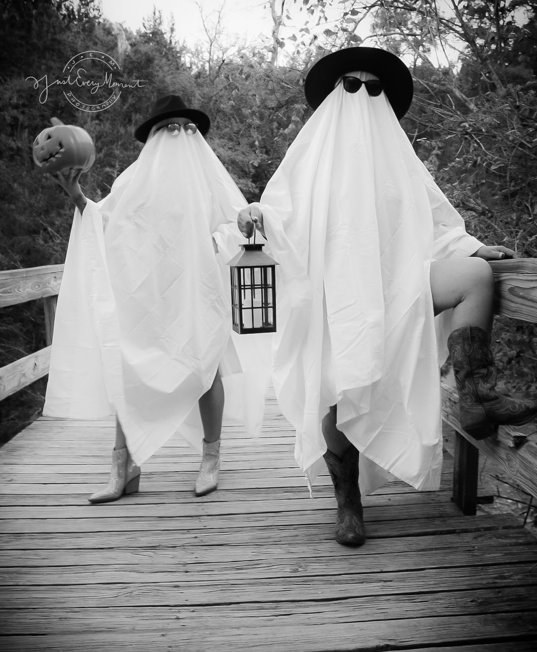 Two figures dressed as ghosts in white sheets and black hats hold a lantern while standing on a wooden bridge.
