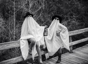 Two people dressed as ghosts in white sheets pose playfully on a wooden bridge in a vintage black and white photograph.