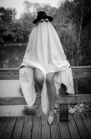 A playful ghost-like figure wearing a black hat sits on a wooden dock by a lake in a black and white photo.