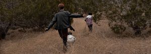 Two people walking together on a dirt hiking trail through brush on a cloudy day in nature.