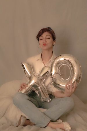 Someone sits cross-legged holding silver XO balloon letters while making a kissy face.