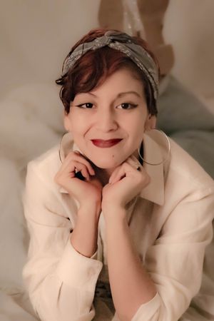 A person wearing a white jacket and headband poses with hands near face in a soft, warm-toned portrait.