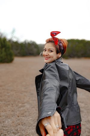 Person wearing a gray raincoat and red bandana holds someone's hand while smiling outdoors on a cloudy day.