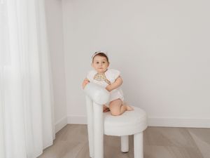 Baby in white onesie sits on modern round white chair against neutral wall and sheer curtain backdrop.