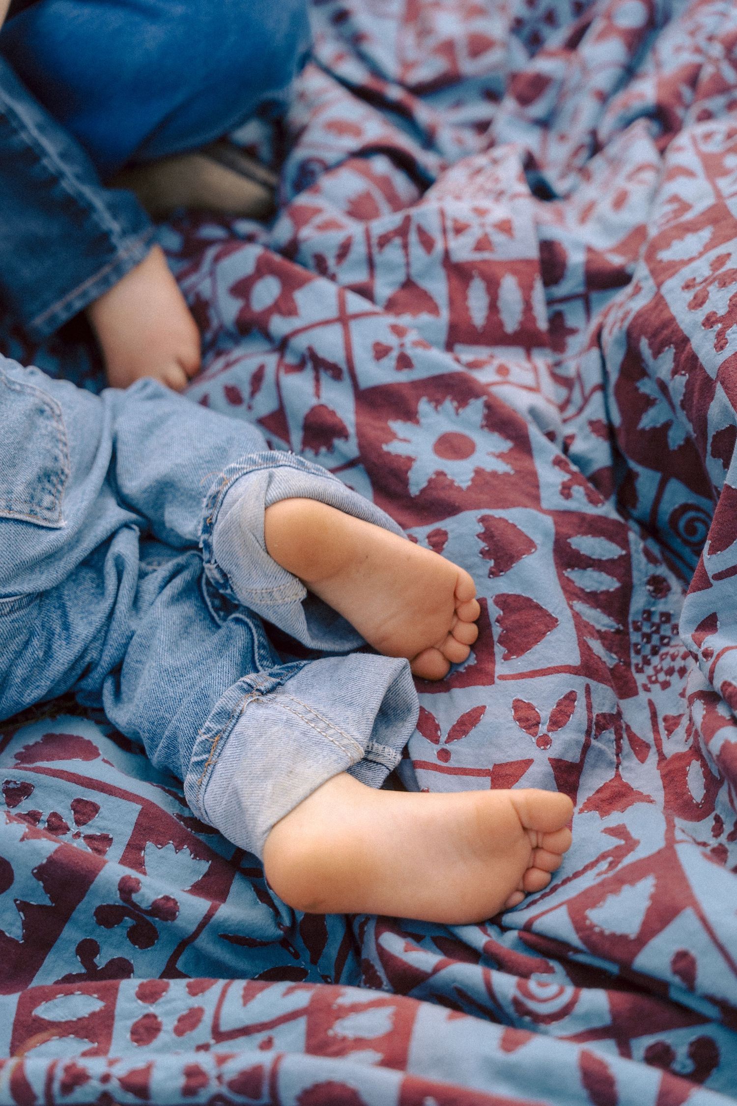 Tiny bare feet of a baby wearing denim overalls resting on a patterned floral blanket.