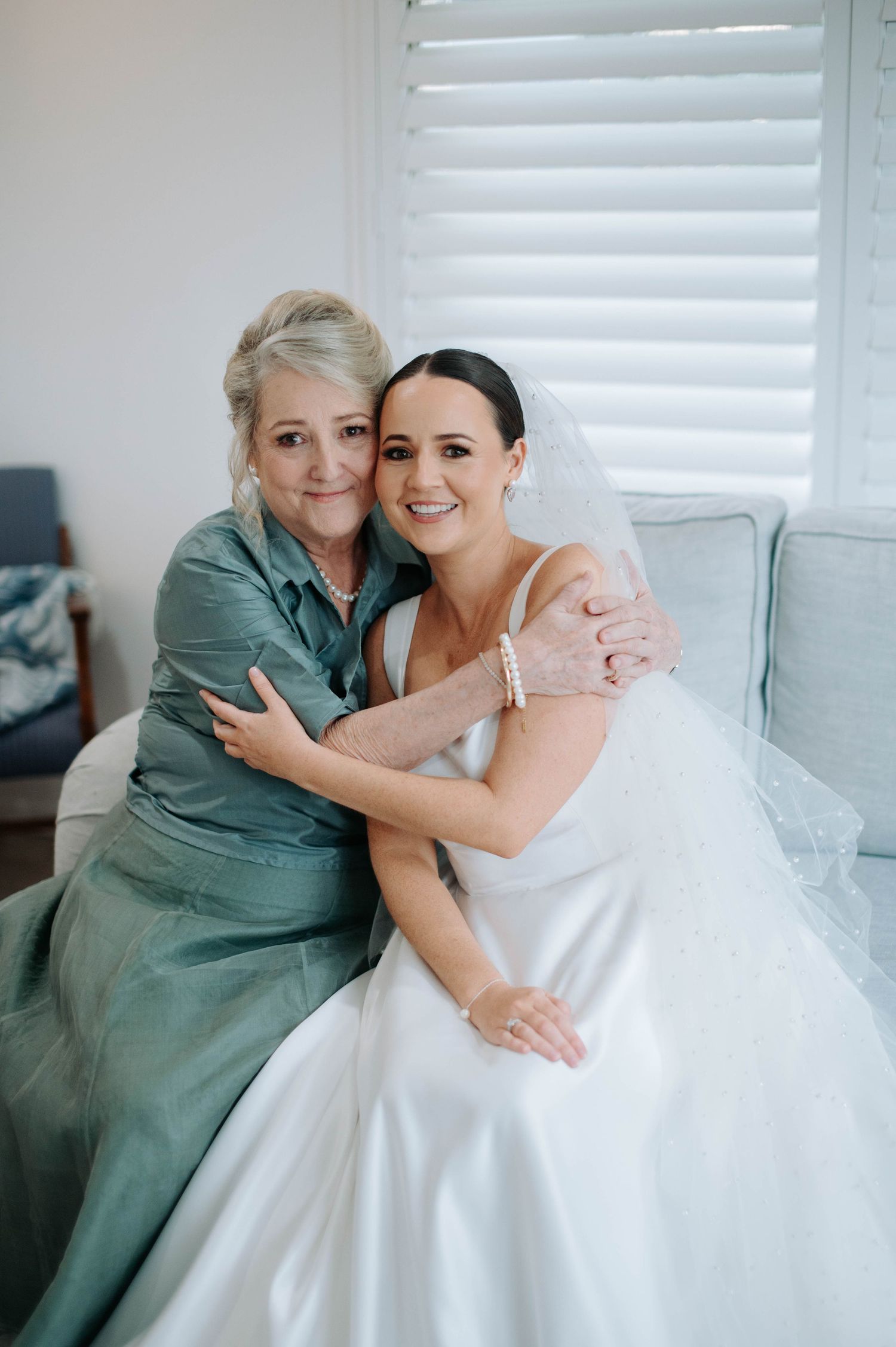 Two women share an emotional embrace on a couch, one in a sage green dress and one in a white wedding gown.