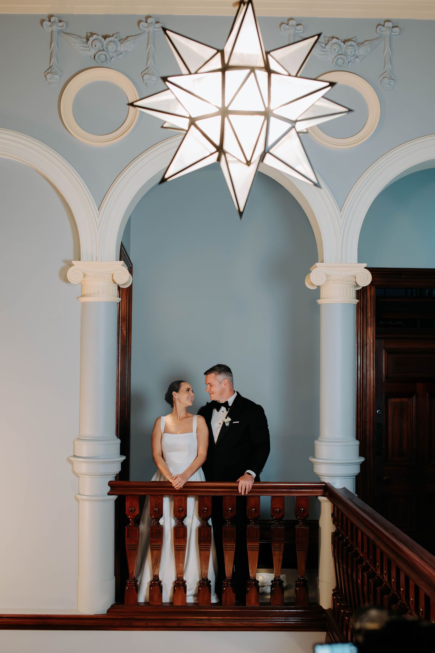 Elegant couple stands beneath ornate white archways and dramatic star-shaped pendant light in historic venue interior.