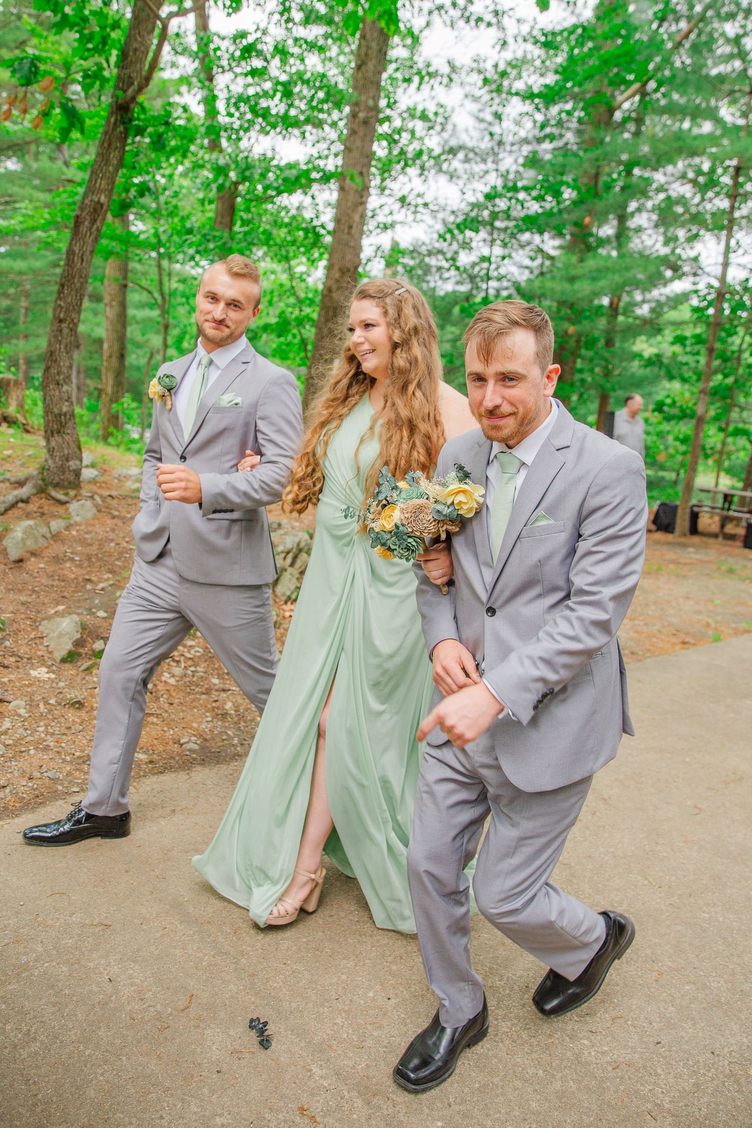 Wedding party having fun on a forest path with individuals in gray suits and a sage green dress smiling and walking.