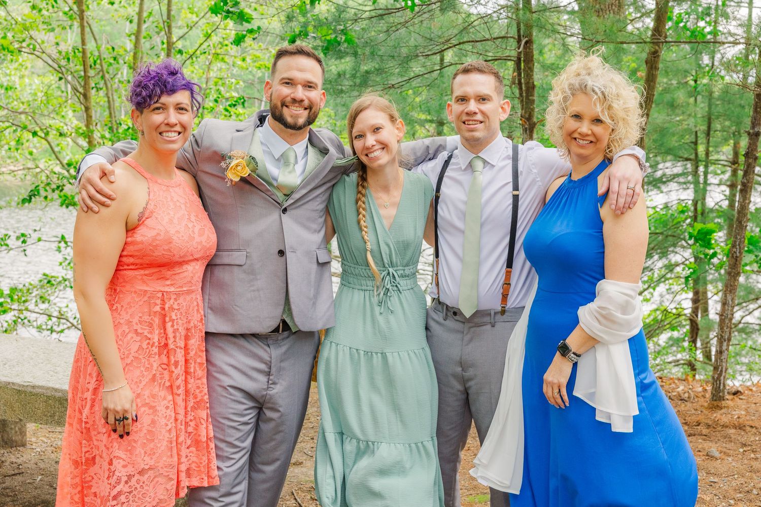 Wedding party members in colorful formal attire smile together in a wooded outdoor setting against green foliage.