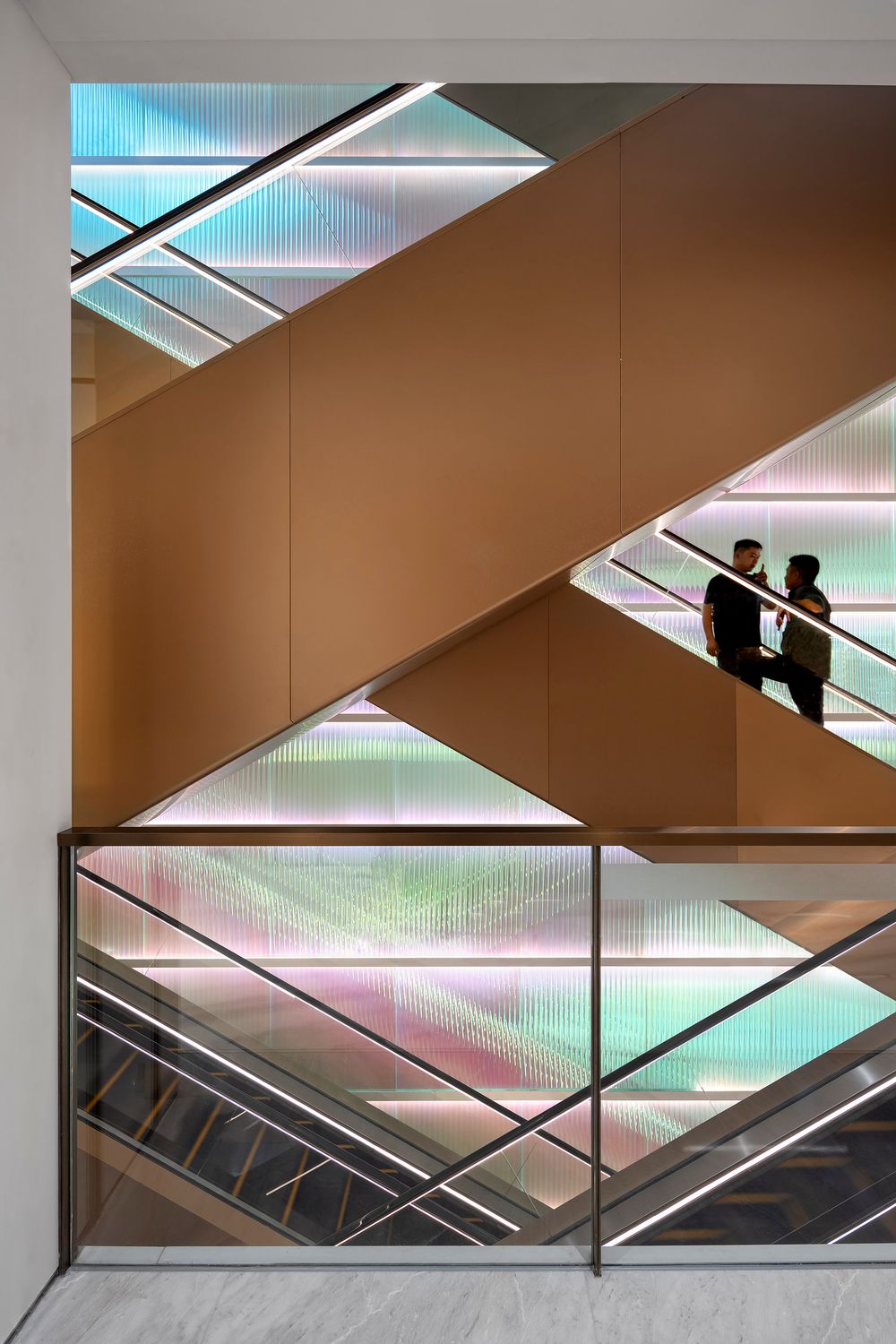 Modern office building interior with geometric escalators and translucent panels creating colorful lighting effects.
