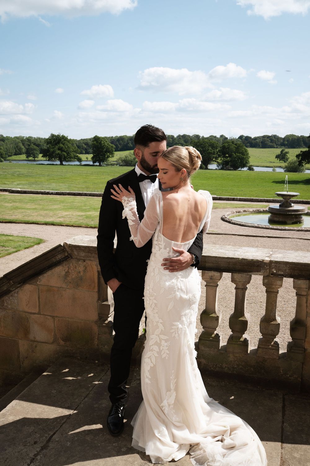 Elegant couple poses on balcony overlooking scenic estate grounds.